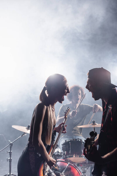 KYIV, UKRAINE - AUGUST 25, 2020: Rock band singers with bass guitars shouting and looking at each other with smoke and blurred drummer on background