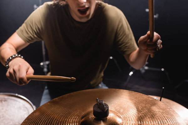 Cropped view of musician with open mouth holding drumsticks and playing cymbal on blurred background