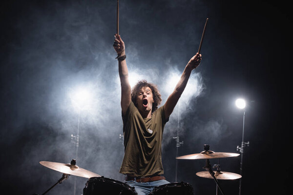 Shouting musician with hands in air sitting at drum kit with smoke and backlit on background