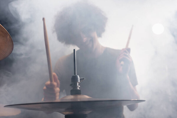 Smiling curly drummer with drumsticks playing drums in smoke