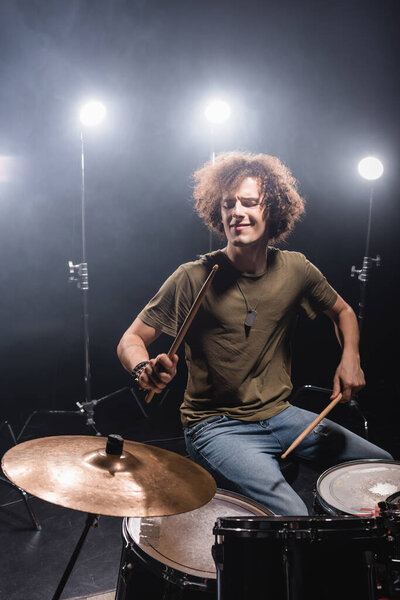 Curly musician playing while sitting at drum kit with drumsticks with back light on background