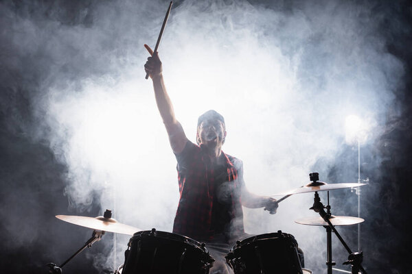 Барабанщик с поднятой рукой, держит барабанные палочки, сидя за барабанной установкой с дымом на черном 