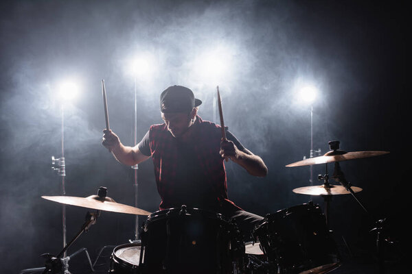 Участник рок-группы играет на барабанах, сидя за барабанной установкой с подсветкой и дымом на заднем плане