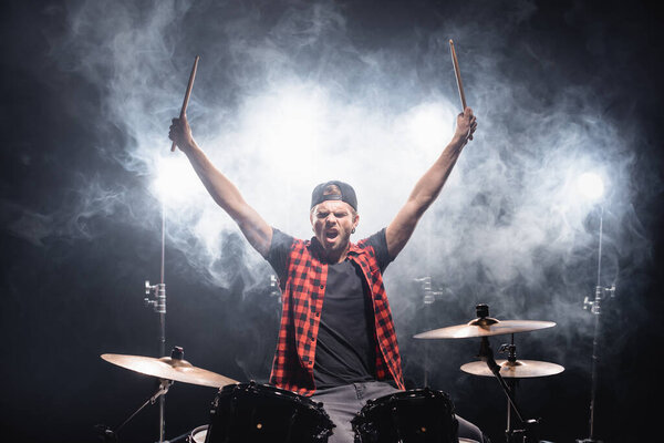 Shouting drummer with hands in air sitting at drum kit with backlit and smoke on black 