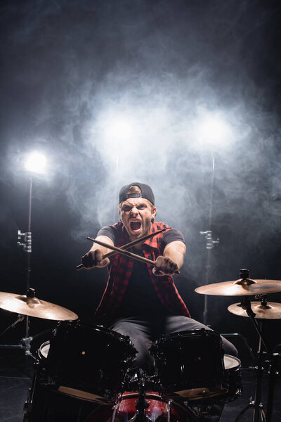Screaming drummer with crossed drumsticks looking at camera, while sitting at drum kit with smoke and backlit on background