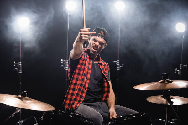 Angry musician showing middle finger, while holding drumsticks and sitting at drum kit with backlit on black