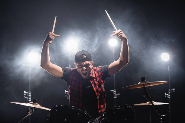 Aggressive drummer with middle fingers looking at camera and holding drumsticks near drum kit with backlit on background