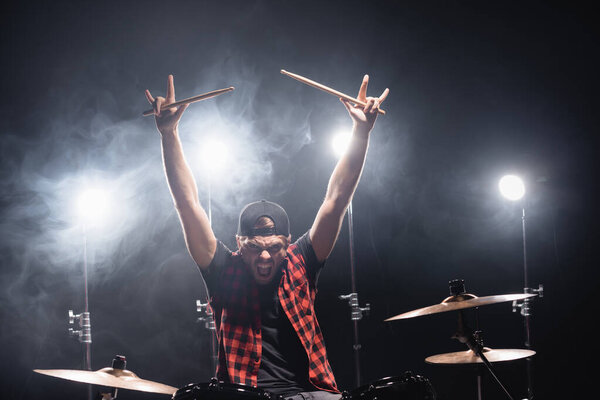 Angry drummer showing rock signs yelling, while sitting at drum kit with backlit on background