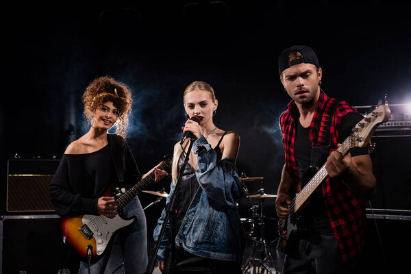 KYIV, UKRAINE - AUGUST 25, 2020: Female singer of rock band standing near musicians with electric guitars on black 