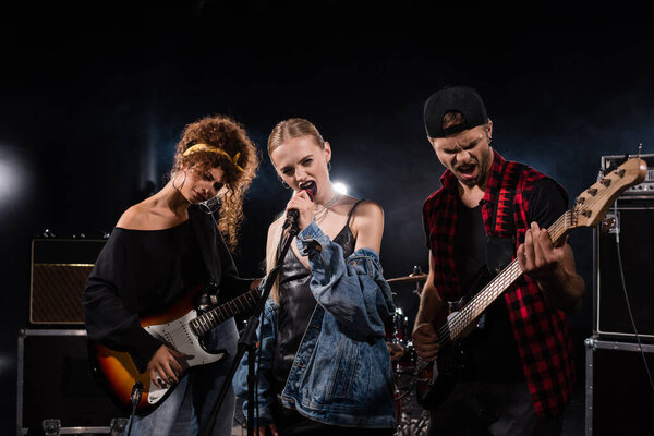 KYIV, UKRAINE - AUGUST 25, 2020: Blonde vocalist of rock band singing near musicians with bass guitars with backlit on black 
