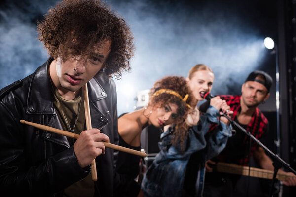 KYIV, UKRAINE - AUGUST 25, 2020: Curly drummer with drumsticks looking at camera with smoke and blurred people on background