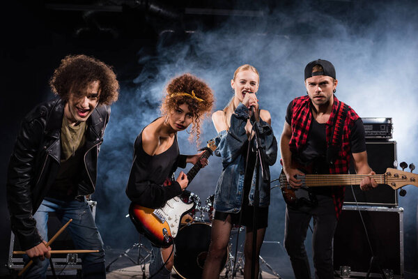 KYIV, UKRAINE - AUGUST 25, 2020: Female vocalist singing, while standing near rock band members with bass guitars and drumsticks, with smoke on black 