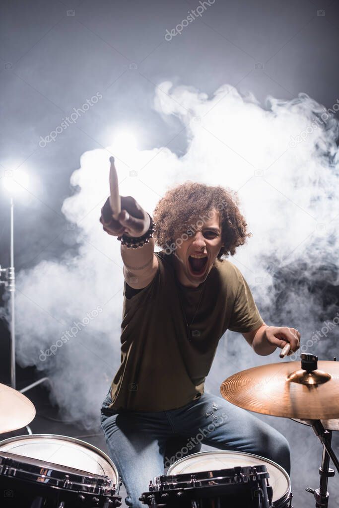 Curly drummer pointing with drumstick while shouting and sitting at drum kit with smoke on background