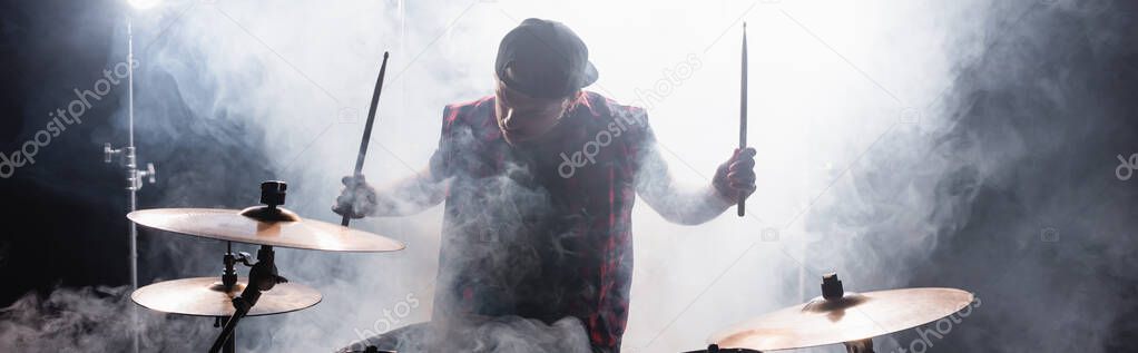 Musician with drumsticks sitting at drum kit with smoke on black background, banner