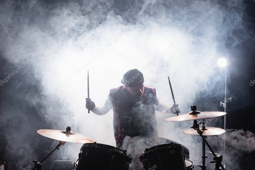 Musician with drumsticks playing while sitting at drum kit with smoke and backlit on background