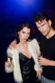 Ernsthafter Mann mit Glas Champagner umarmt Freundin in Kleid und weißer Jacke mit Hintergrundbeleuchtung auf Schwarz