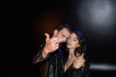 Kızgın adamın orta parmağını göstermesi seksi bir kadının yanında duruyor. Siyah üzerine taş bir jest, bulanık arka planda.