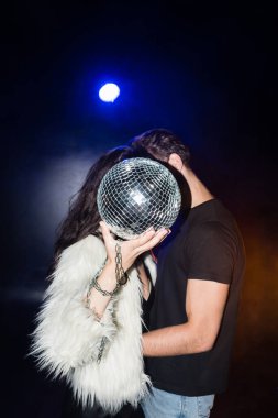 Beyaz ceketli kıvırcık kadın disko topu tutarken, siyah arka ışıklandırmalı erkek arkadaşıyla kucaklaşıyor.