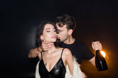 Şampanya şişeli tutkulu bir adam siyah arka ışıklandırmalı seksi bir kadının boynuna dokunuyor.
