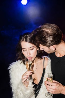 Gömlekli bir adam, elinde bir bardak şampanya, arkasında siyah ışık olan baştan çıkarıcı esmer bir kadına bakıyor.