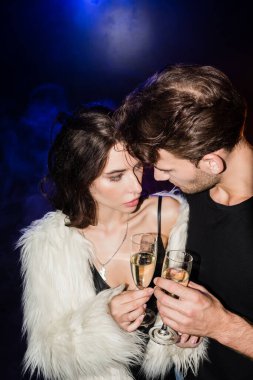Erkek ve baştan çıkarıcı kadın şampanya kadehleriyle kadeh kaldırırken birbirlerine arka planda siyah ışıklarla bakıyorlar.
