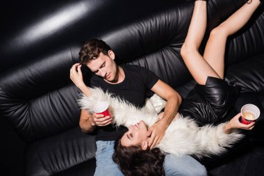Plastik bardaklı bir adam, gece kulübünde bacaklarıyla kanepede uzanmış kadına dokunuyor.