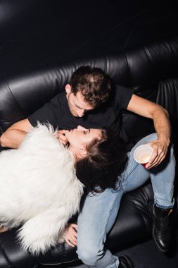 Kanepede ayağı olan bir adamın yüksek açılı görüntüsü, gece kulübünde plastik kahve tutarken kadının çenesine dokunması,
