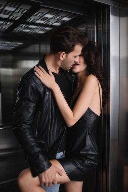 Deri ceketli bir adam asansörde siyah elbiseli, baştan çıkarıcı bir kadının bacağını öpüyor.