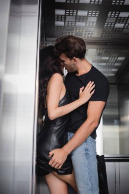 Siyah tişörtlü ve kot pantolonlu bir adam asansörde kıvırcık bir kadının poposuna dokunuyor.