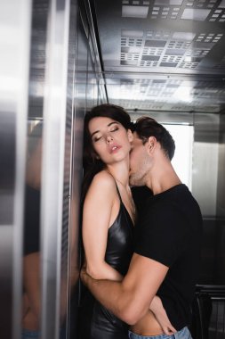 Siyah tişörtlü tutkulu bir adam asansörde boynunu öperken gözleri kapalı seksi bir kadına sarılıyor.