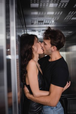 Siyah elbiseli tutkulu bir çift asansörde sarılıp öpüşüyor.
