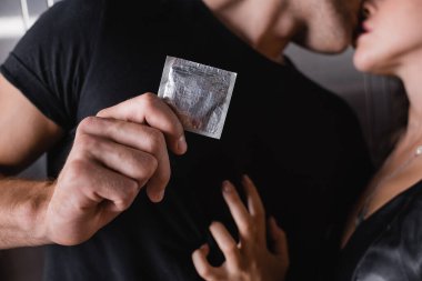 Siyah tişörtlü, tutkulu bir adamın arka planda bir kadını öperken prezervatif göstermesi.