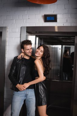 Kıvırcık saçlı neşeli kadın asansör girişinin yanında deri ceketli, gülümseyen adamı kucaklıyor.