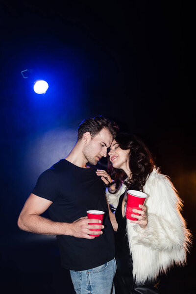 Smiling woman with closed eyes touching boyfriend shirt, while holding plastic cup with backlit on black