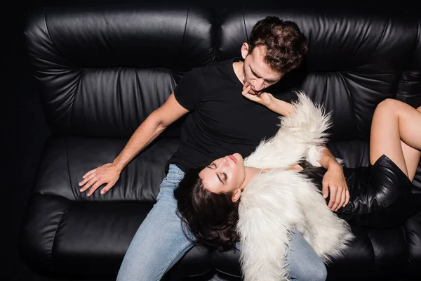 Passionate brunette woman in white jacket touching boyfriend chin, while lying on sofa in nightclub