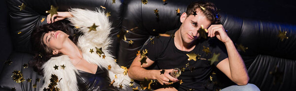 Woman in white jacket and dress lying near man with glass of champagne sitting on sofa, while confetti falling in nightclub, banner
