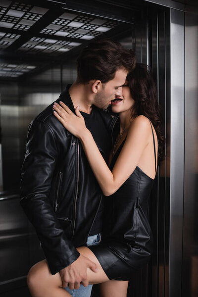 Мужчина в кожаной куртке целуется и держит ногу соблазнительной женщины в черном платье в лифте