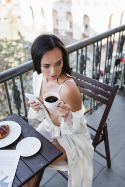 Beyaz bornozlu güzel esmer kadın elinde akıllı telefonla balkonda kahve içiyor.