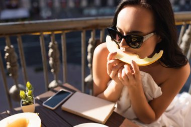 brunette woman in sunglasses and covered in white sheet eating melon on balcony clipart