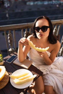 Güneş gözlüklü, beyaz çarşaflı ve balkonda kavun yiyen seksi esmer kadın.
