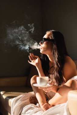 seksi esmer kadın beyaz çarşafa sarılmış sigara içiyor ve kahve içiyor.