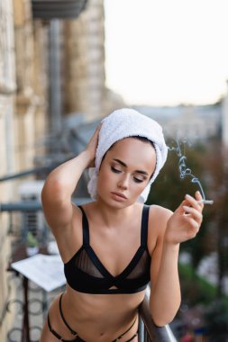Siyah iç çamaşırlı, kafasında havluyla balkonda sigara içen seksi genç bir kadın.