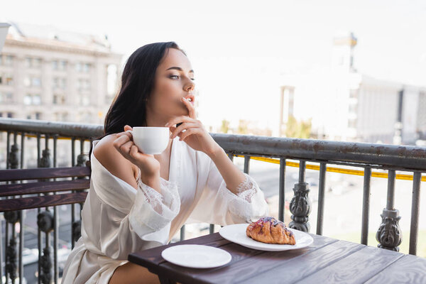 brunette woman in white robe drinking coffee and holding fingers near mouth