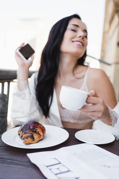 白い服を着た美しいブルネットの女性がスマートフォンを持っていてバルコニーで朝食を取っています — ストック写真