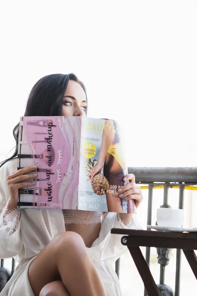 身穿白袍的黑发女人坐在阳台上看杂志 — 图库照片