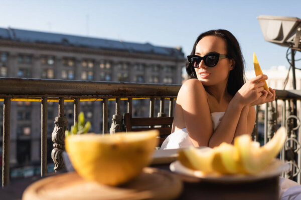 сексуальная брюнетка в солнечных очках и покрытая белым полотном, поедающая дыню на балконе