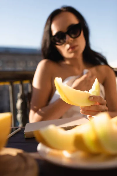 sexy brunette woman in sunglasses and covered in white sheet eating melon on balcony