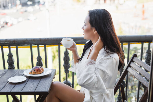 brunette woman in white robe drinking tea and sitting on balcony