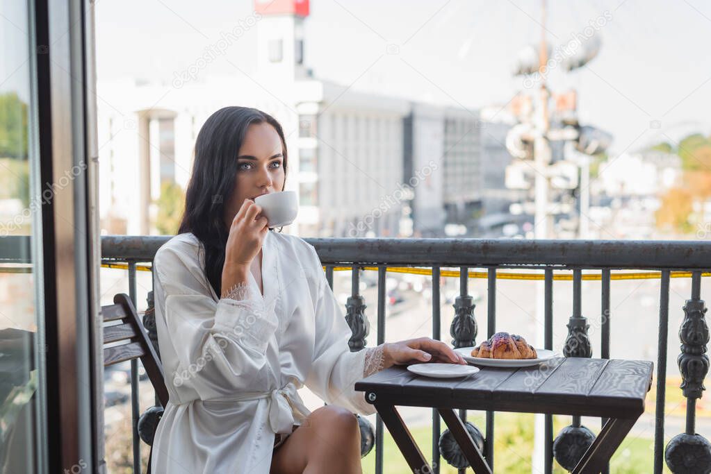 brunette woman in white robe drinking tea and sitting on balcony