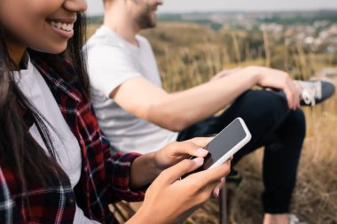 Kamp sırasında arka planda bulanık bir şekilde akıllı telefon kullanan gülümseyen Afro-Amerikan kadın görüntüsü. 